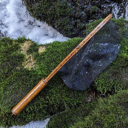 A bamboo fly fishing net lying on a rock in the water on the river in Western North Carolina. The rubber net basket is smooth and shiny and protects the fish for catch and release. The handle is flame cured for strength and durability and there is copper stitching and a copper keeper ring to attach your net keeper to.