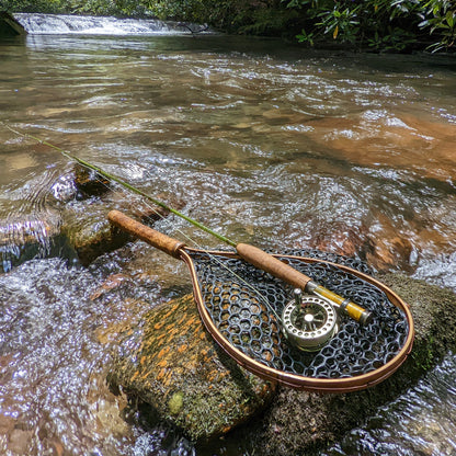 A bamboo fly fishing net lying on a rock in the water on the river in Western North Carolina. The rubber net basket is smooth and shiny and protects the fish for catch and release. The handle is flame cured for strength and durability and there is copper stitching and a copper keeper ring to attach your net keeper to.