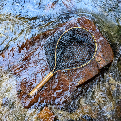 A bamboo fly fishing net lying on a rock in the water on the river in Western North Carolina. The rubber net basket is smooth and shiny and protects the fish for catch and release. The handle is flame cured for strength and durability and there is copper stitching and a copper keeper ring to attach your net keeper to.